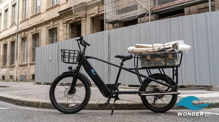 Le vélo cargo Wonder Eco chargé d'une caisse à outils et de bâches roulées, devant un immeuble en rénovation.